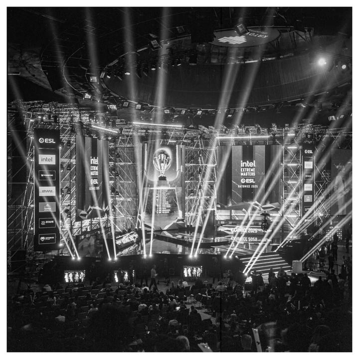 A large esports stage with bright spotlights illuminates a trophy and banners in front of an audience.