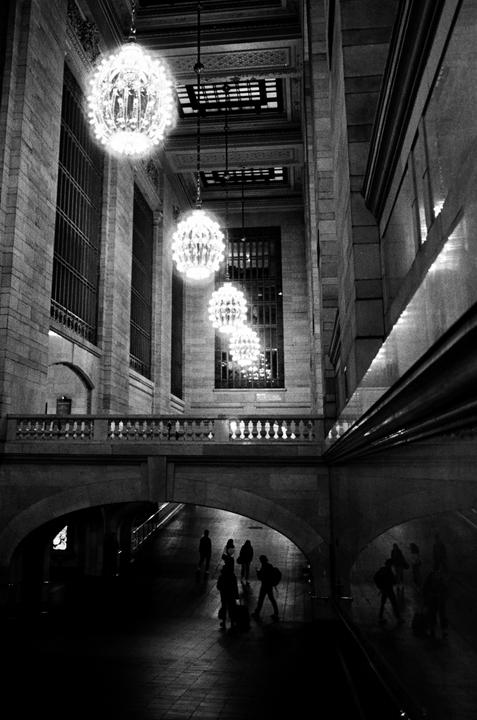 A black and white photograph shows a grand hall with ornate chandeliers and people walking below an arched bridge.