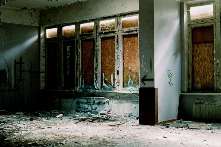 Sunlight streams into a derelict room with boarded-up windows and debris on the floor.