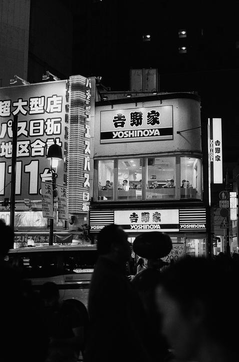 A street at night is filled with neon signs and people, with a Yoshinoya restaurant visible.