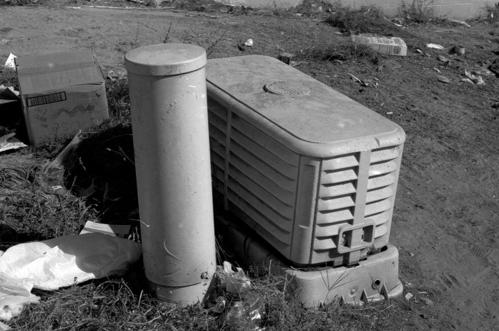 A cylindrical post stands next to a rectangular utility box on a grassy, dirt slope with scattered trash.