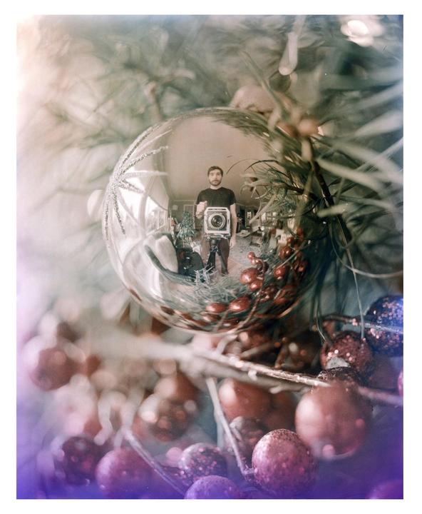A man holding a camera is reflected in a Christmas ornament hanging on a Christmas tree.