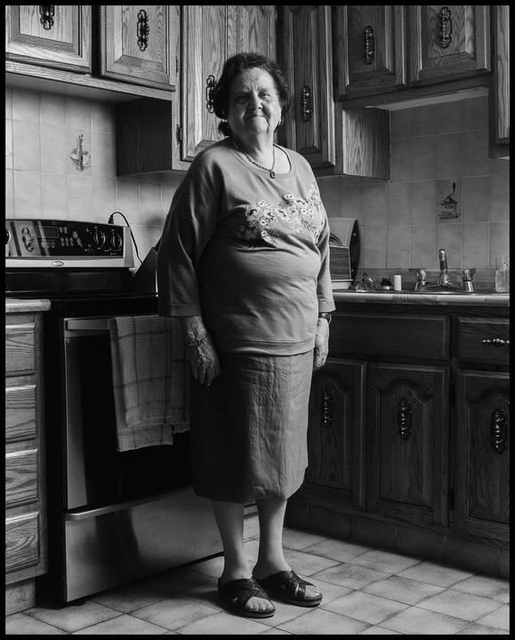 A woman stands in a kitchen with wooden cabinets and a tiled floor.