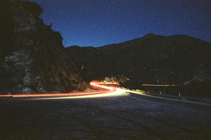 Light trails from vehicles illuminate a winding road at night, with mountains and a starry sky in the background.