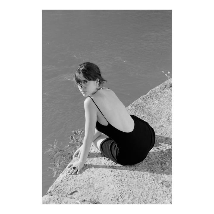 A woman in a black dress crouches on a stone ledge overlooking water.