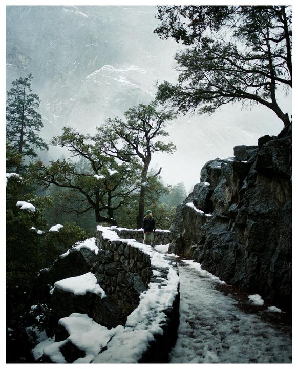 A woman stands on a snowy stone path next to a rocky cliff with trees and mountains in the background.