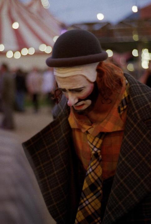 A clown in a bowler hat, plaid jacket, and plaid tie looks down with a sad expression.
