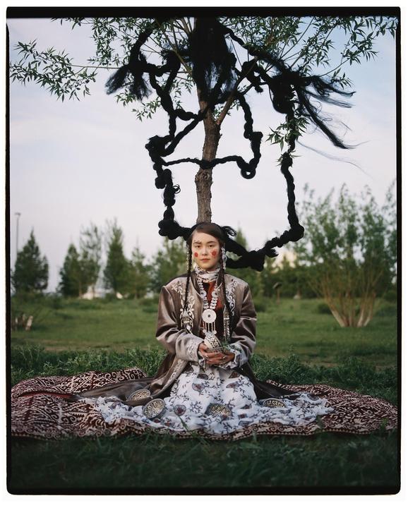 A woman in traditional clothing with elaborate braids sits on a patterned blanket under a tree with braids woven into its branches.