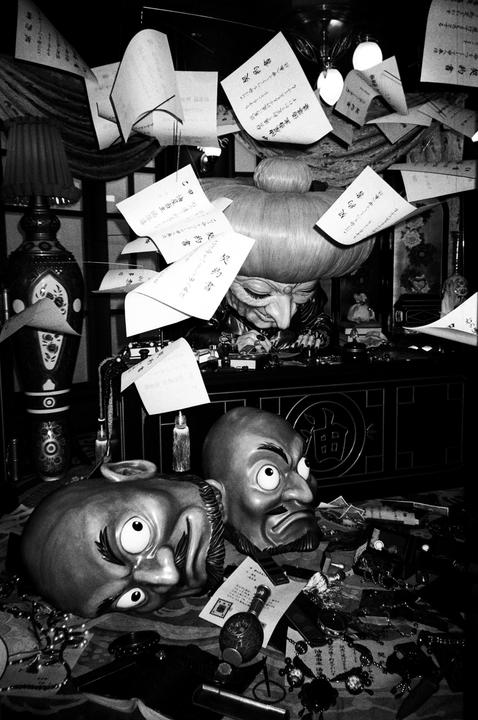 Two masks and a figure with a large head are surrounded by papers and various objects on a table.