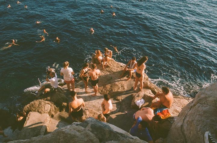 A group of people are swimming in the ocean and relaxing on rocks by the water.