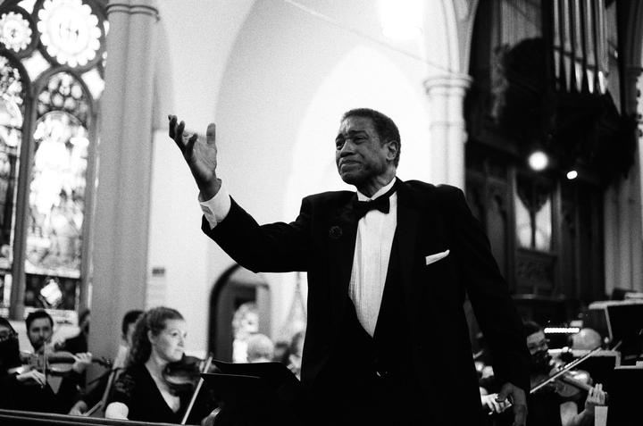 A man in a tuxedo conducts an orchestra in a large hall with stained glass windows.