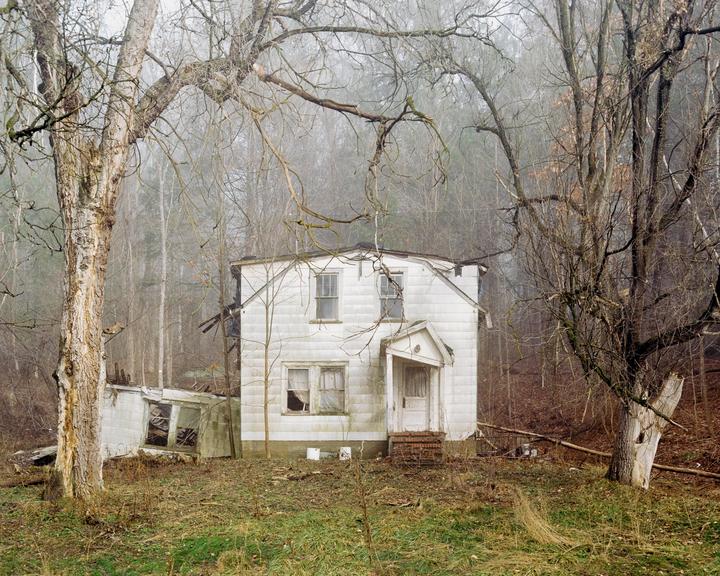 An abandoned white house with broken windows stands among bare trees in a foggy forest.