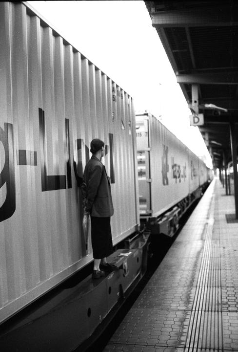 A woman stands on a train platform next to a long freight train.