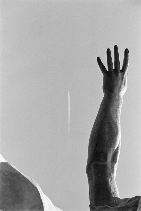 A statue's arm and hand reach towards the sky, with an airplane contrail visible in the distance.