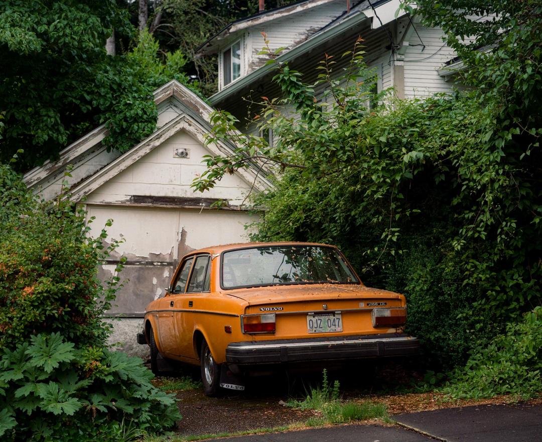 An orange car is parked in front of an overgrown garage with peeling paint.