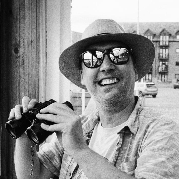 A man wearing a hat and sunglasses smiles while holding binoculars.