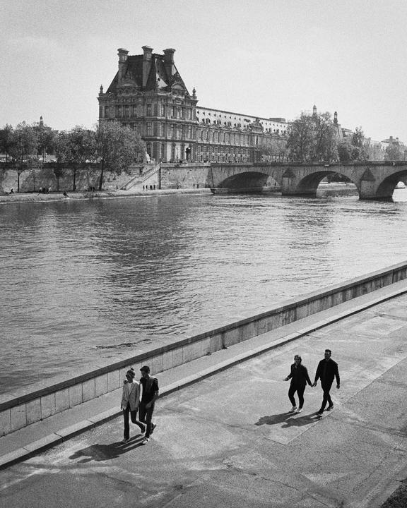 Two couples walk along a river with a large building and bridge in the background.