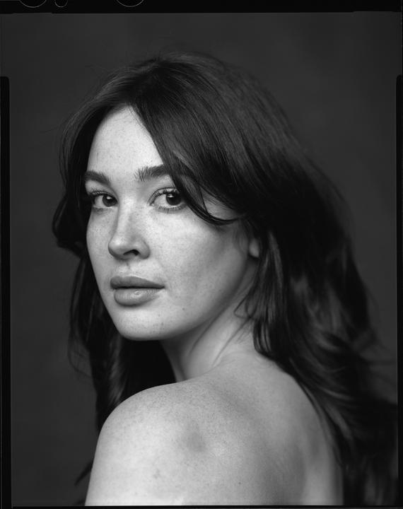 A woman with long hair and freckles looks over her shoulder at the viewer.