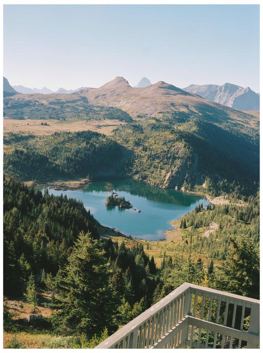 A mountain lake with a small island is surrounded by a forest and mountains under a clear sky.