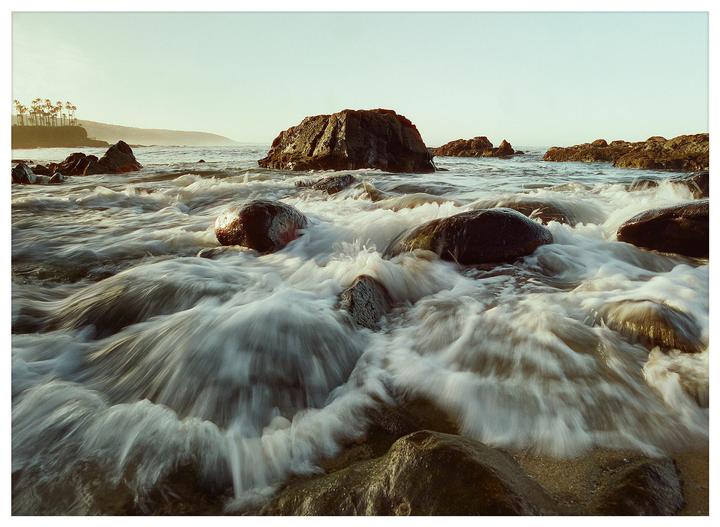 Waves crash over rocks on a beach with palm trees visible in the distance under a bright sky.