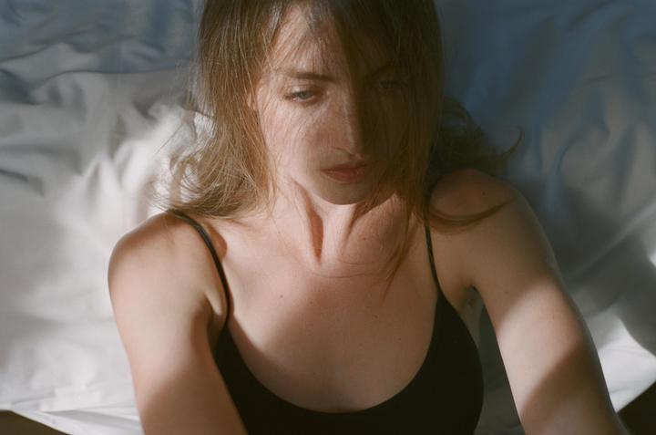 A woman with long hair and a black top lies on a white sheet with shadows on her face and body.