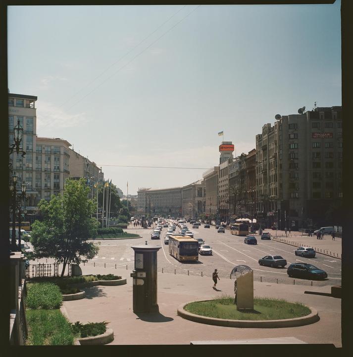A wide street with cars and buses is lined with buildings and trees under a clear sky.