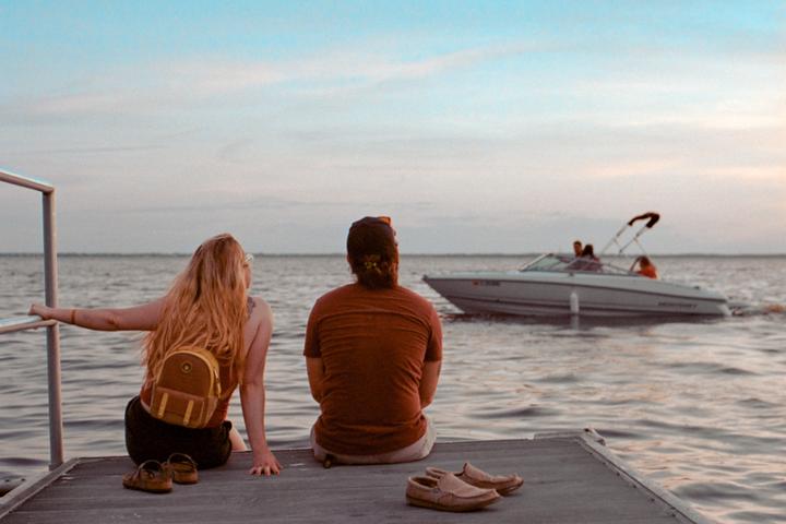 A woman and a man sit on a dock looking out at a boat on the water.