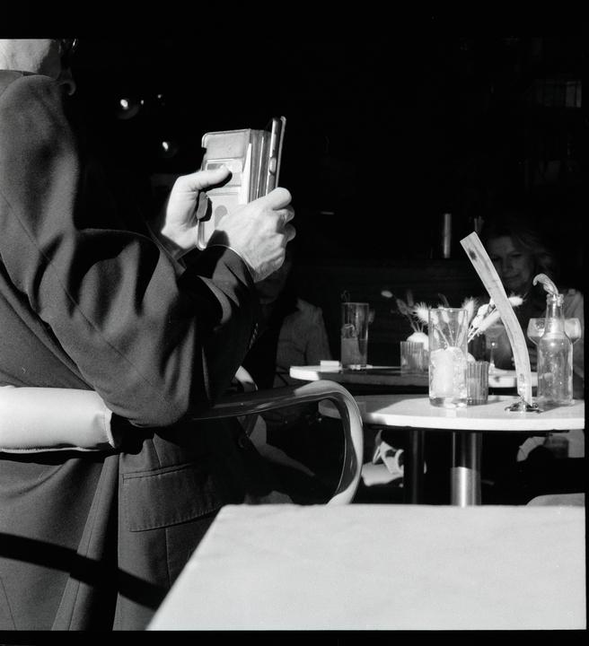 A man holds a phone at a table with drinks and a woman in the background.