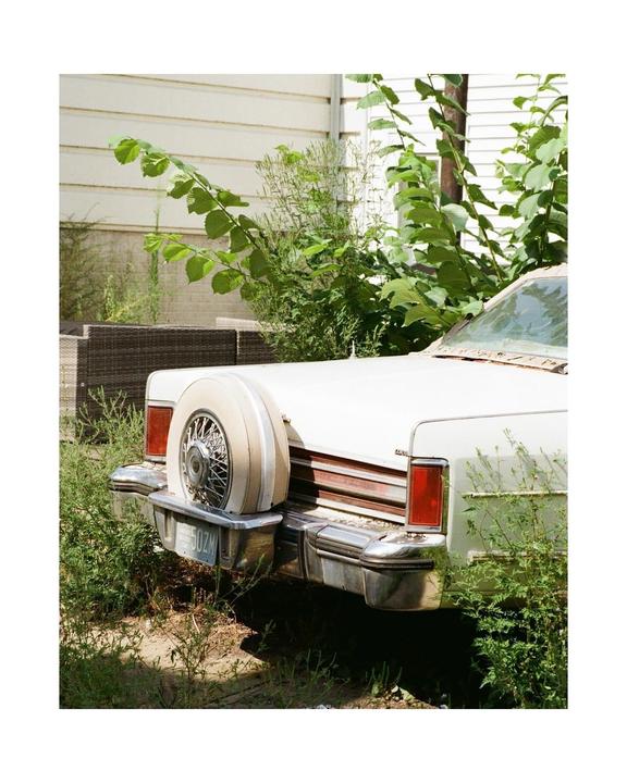 A white vintage car with a spare tire on the back is parked in an overgrown yard next to a house.