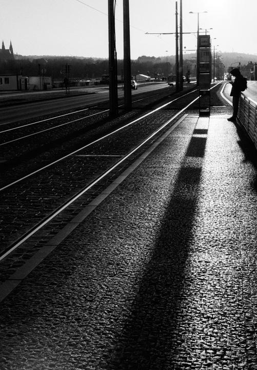 A person stands on a tram platform next to tram tracks, with a road and buildings in the background.