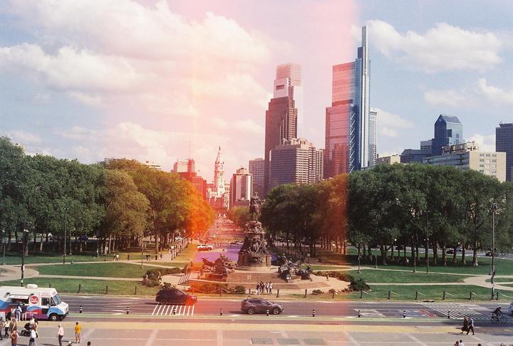 A wide view of a city park with a monument in the center, surrounded by trees and buildings under a cloudy sky.