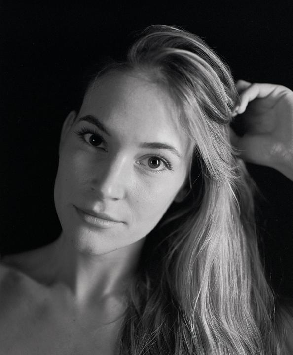 A woman with long hair looks directly at the viewer, touching her hair with one hand.