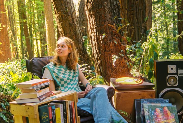 A woman sits in an armchair in a forest with a record player and books.