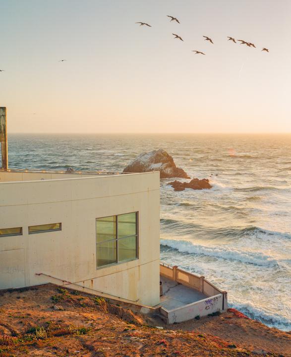 A flock of birds flies over a building on a cliff overlooking the ocean at sunset.