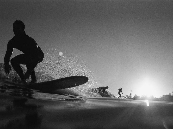 A surfer rides a wave with the sun low on the horizon, creating a strong backlight.