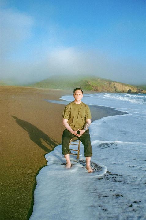 A man sits on a chair in the surf on a beach with a hazy mountain in the background.