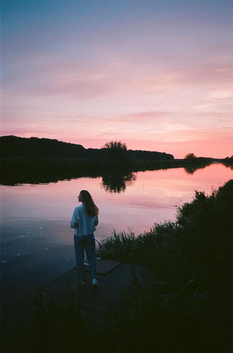 A woman stands on a dock looking out at a river with a pink and purple sunset.