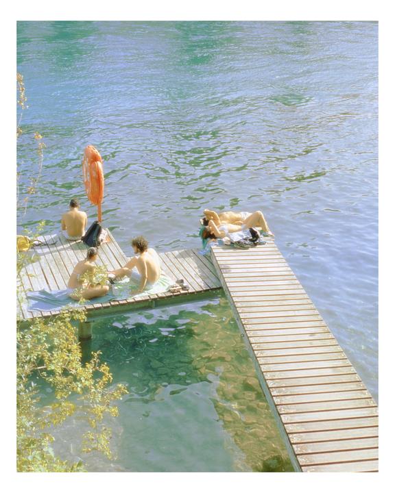 Four people are relaxing on a wooden dock next to clear blue water.