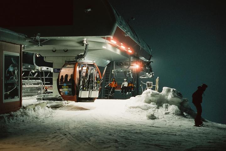 A snowboarder stands on a snowy slope next to a ski lift station with a gondola and chairlift at night.
