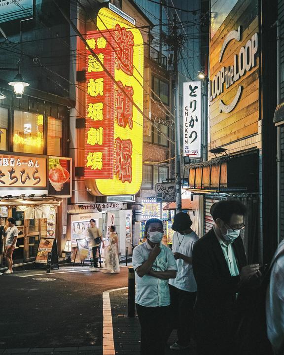 People walk along a narrow street lined with brightly lit signs and buildings at night.