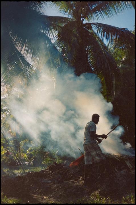 A man stands in a cloud of smoke, holding a long stick, with palm trees in the background.