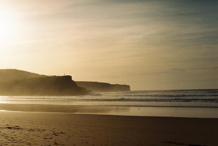 A lone person stands on a cliff overlooking a beach with waves crashing on the shore under a hazy sky.