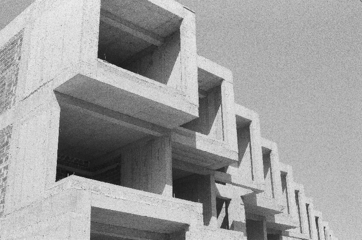 A concrete building with many square openings is shown from a low angle against a bright sky.