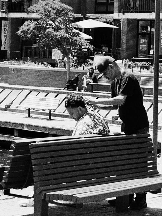 A man wearing a baseball cap is cutting another man's hair on a wooden bench next to a body of water.