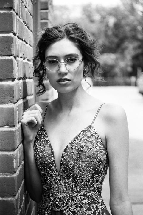 A woman in a formal dress and glasses leans against a brick wall.