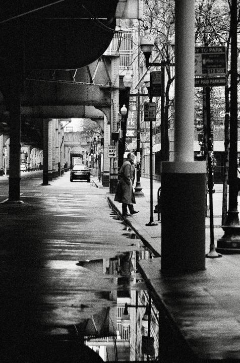 A man stands on a wet sidewalk next to a street with a car and an elevated train track overhead.