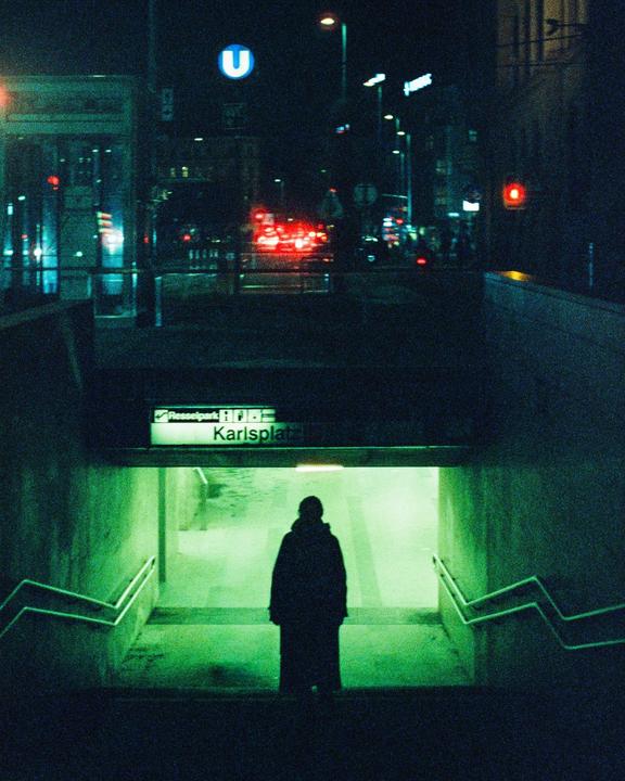 A person stands at the top of a subway entrance with a street and traffic visible in the background at night.
