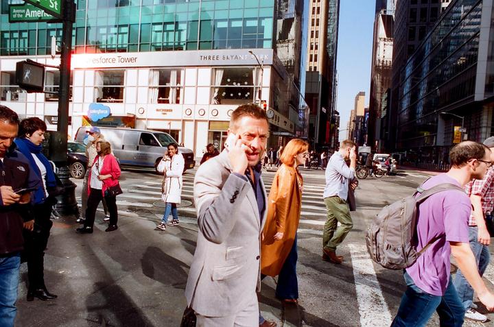 A man in a suit talks on a phone while crossing a street with other people and tall buildings in the background.