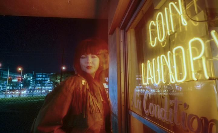 A woman with bangs and a fringed jacket stands next to a neon "Coin Laundry" sign at night.