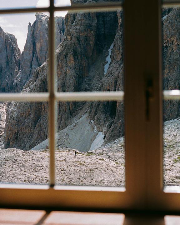 A man stands on a rocky mountain slope, viewed through a window.